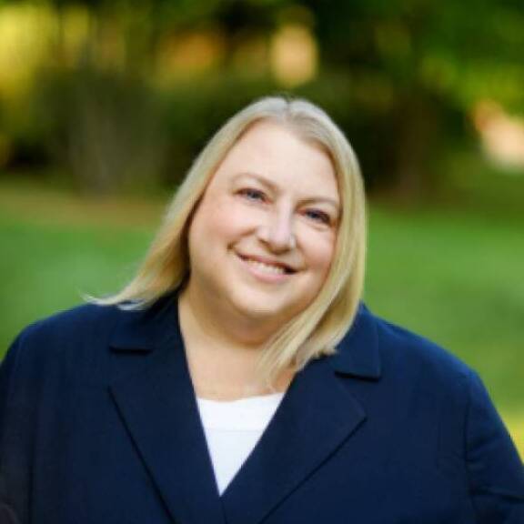 Smiling blonde woman standing in green grass wearing a a black blazer and white shirt